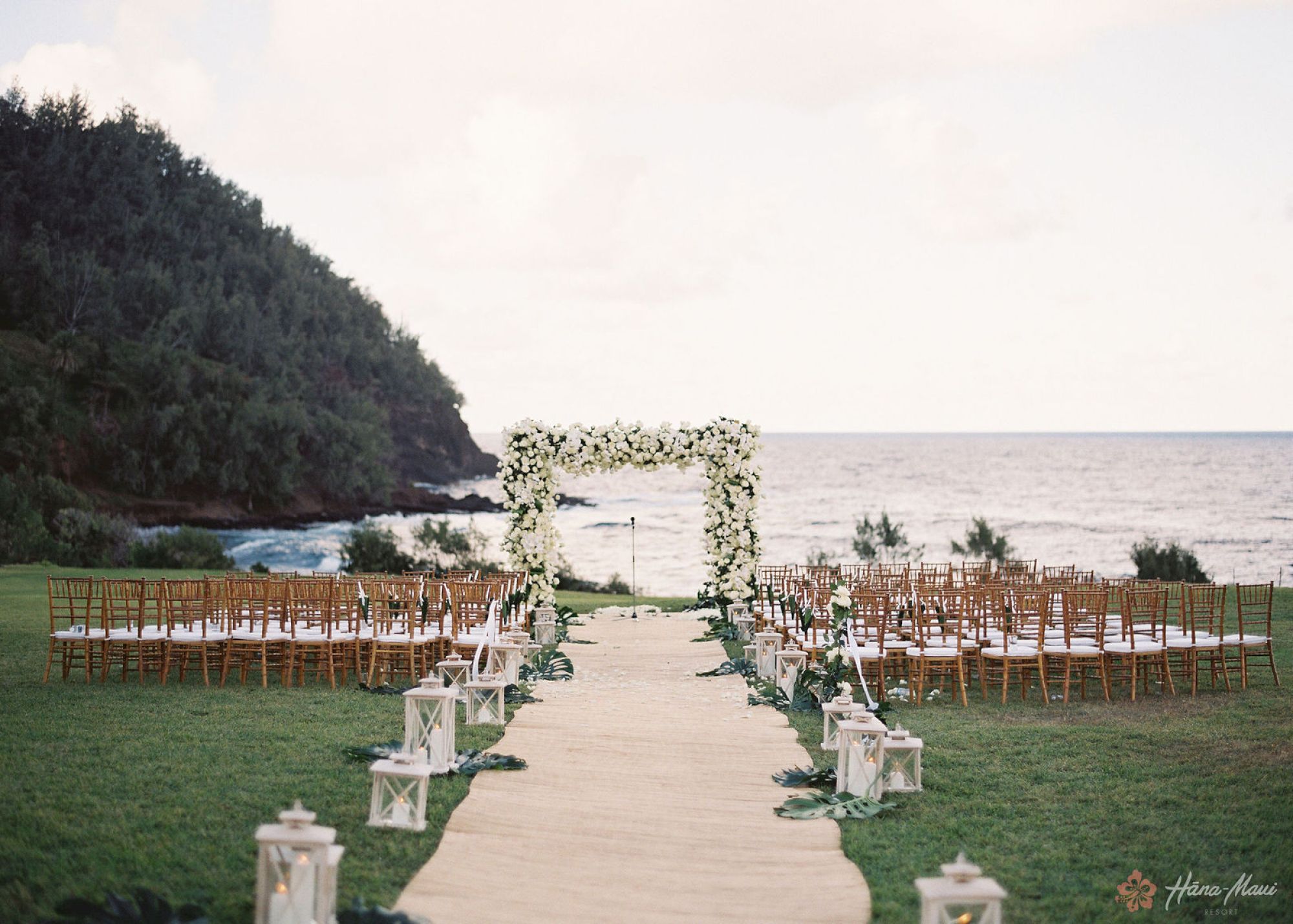 An outdoor wedding setup with a floral arch overlooking the ocean. Rows of chairs are aligned on either side of a white aisle runner, ending the sentence.