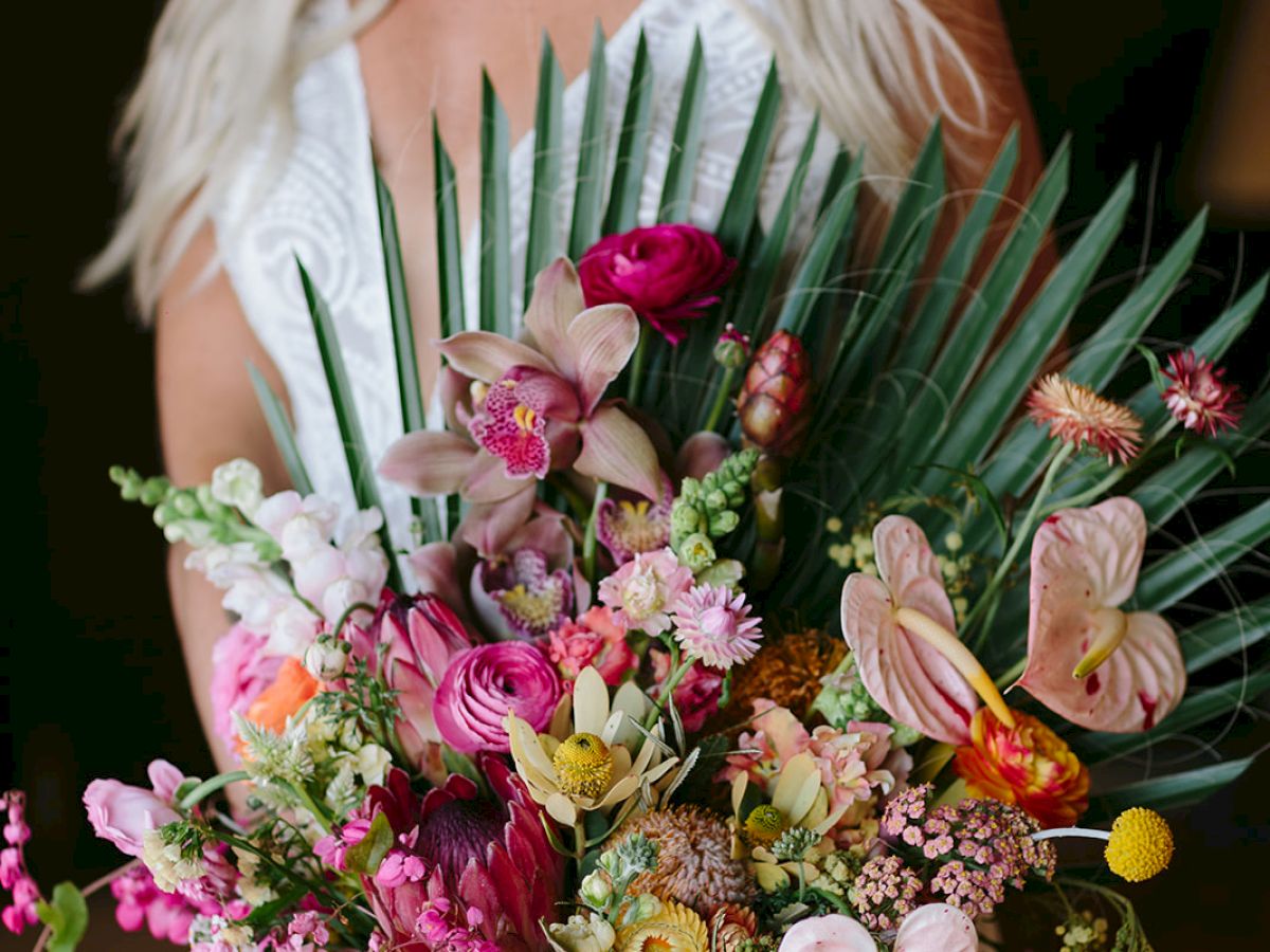 A woman in a white dress holds an elaborate bouquet of colorful flowers and greenery, including orchids and palm leaves.