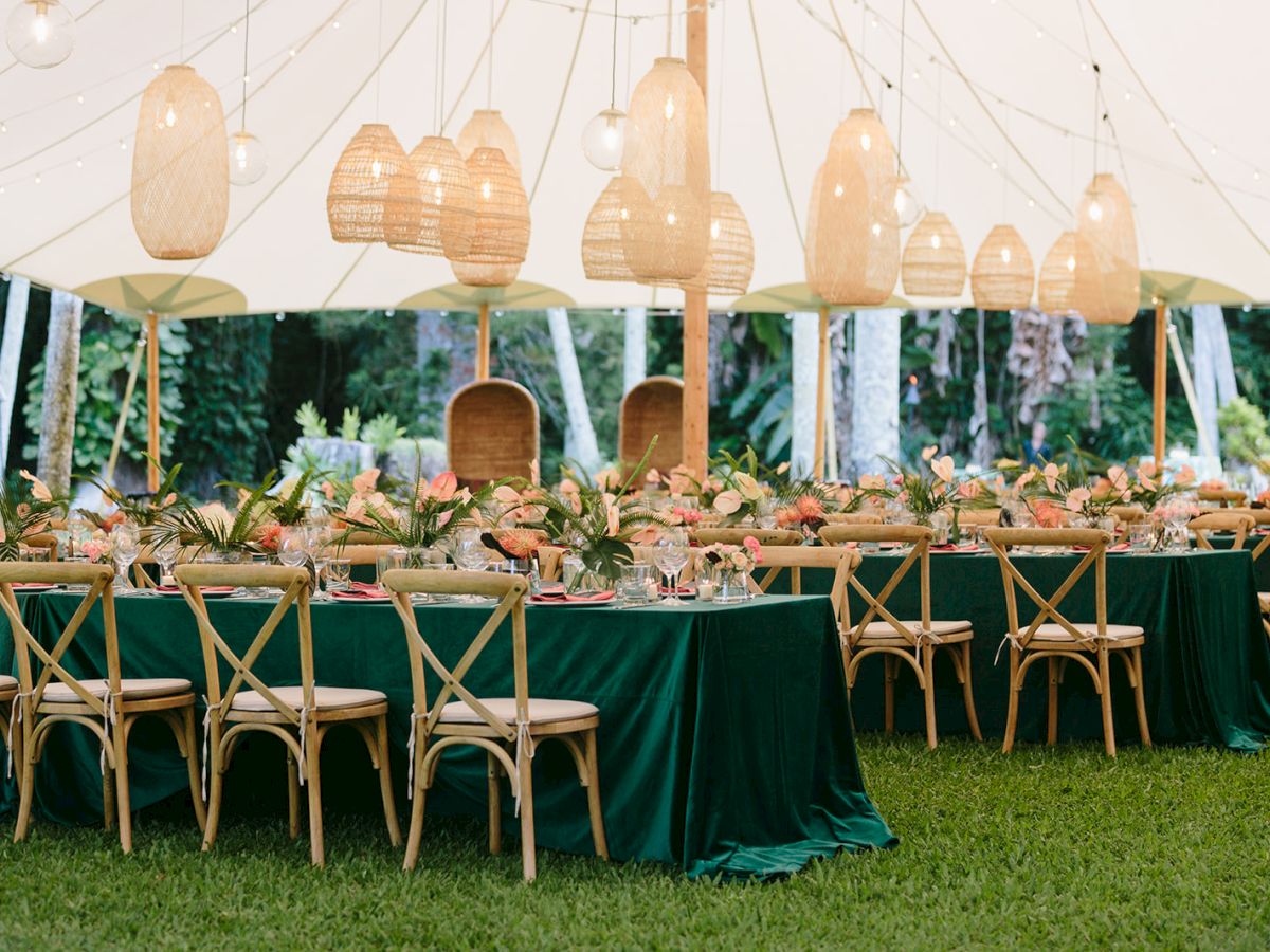 An outdoor event setup features long tables with green tablecloths, wooden chairs, and overhead lantern decorations under a canopy tent.