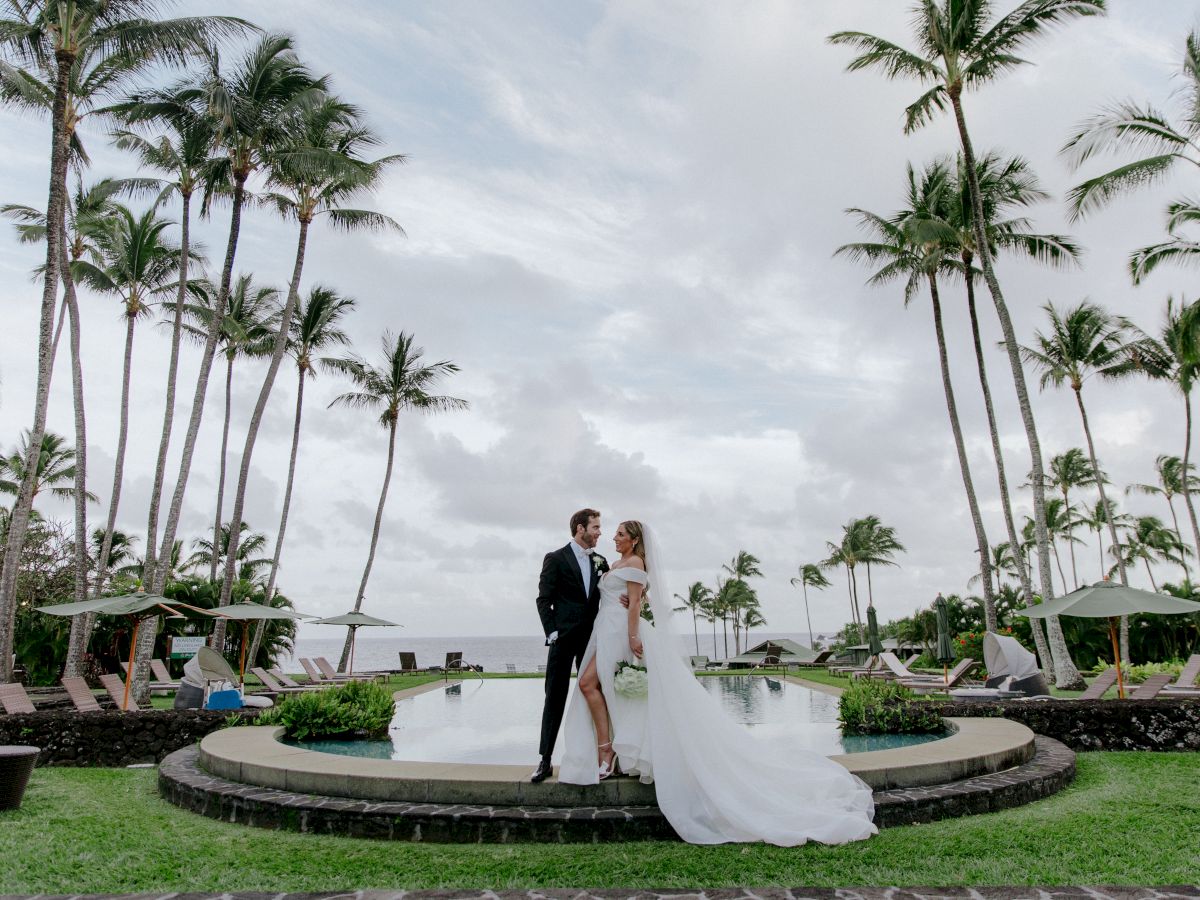 A bride and groom stand together near a circular pool in a tropical setting with tall palm trees surrounding them, with a cloudy sky above.