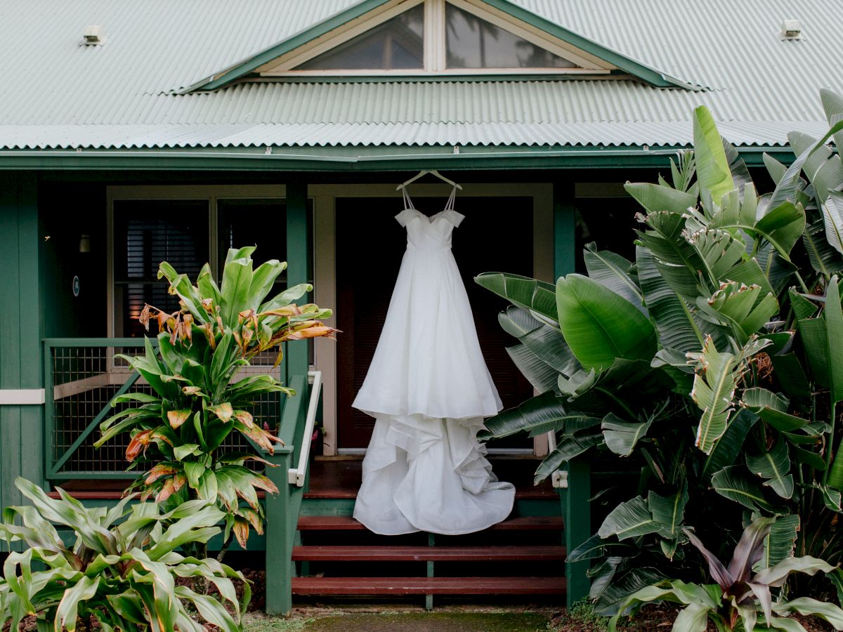 A white wedding dress hangs on a porch next to lush green plants and a green house, creating a serene and picturesque scene.