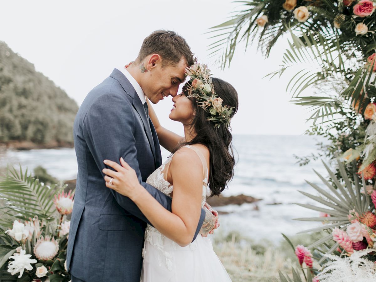 A couple embraced forehead to forehead, in wedding attire, surrounded by tropical flowers and foliage near a serene ocean.