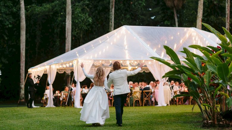 A couple is walking hand in hand towards a decorated outdoor event tent with string lights, surrounded by lush greenery.