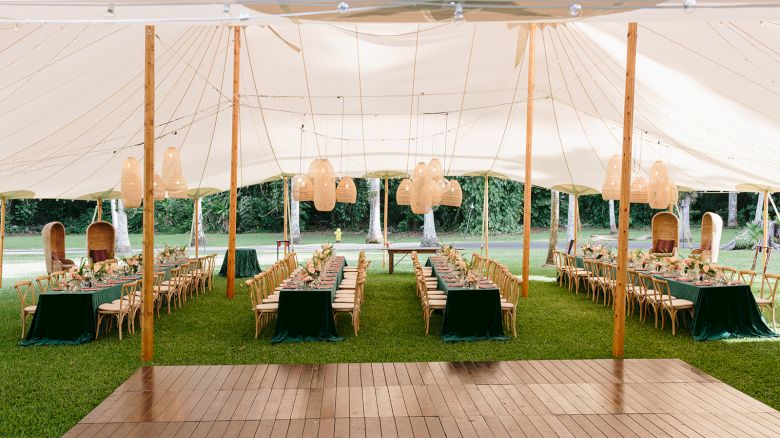 An outdoor event tent set up with long dining tables, chairs, and hanging lanterns over a grass lawn, with wooden flooring in the foreground.