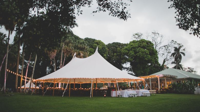 A spacious outdoor event tent with string lights is set up on a grassy area, next to some trees and a building, creating a festive atmosphere.