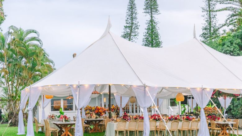 The image shows an outdoor event tent with draped white fabric and decorated tables set for a gathering in a garden setting, surrounded by greenery.