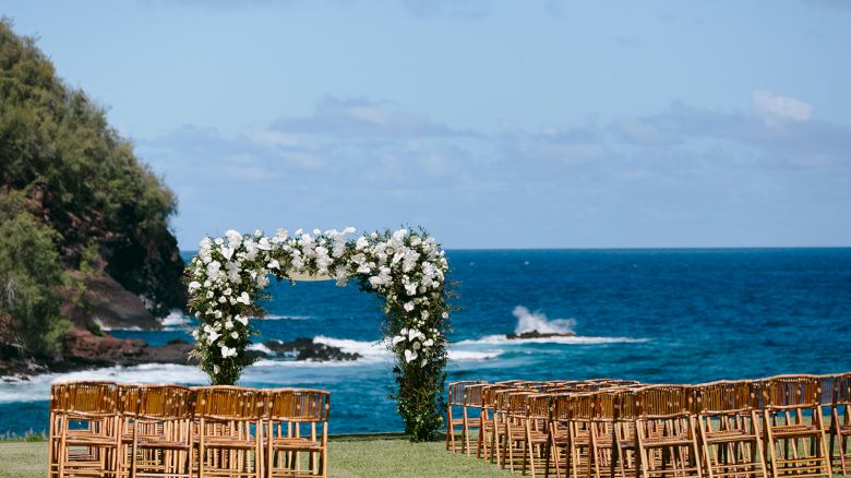 An outdoor wedding setup with wooden chairs and a floral arch overlooking the ocean against a backdrop of cliffs and blue sky, ending the sentence.