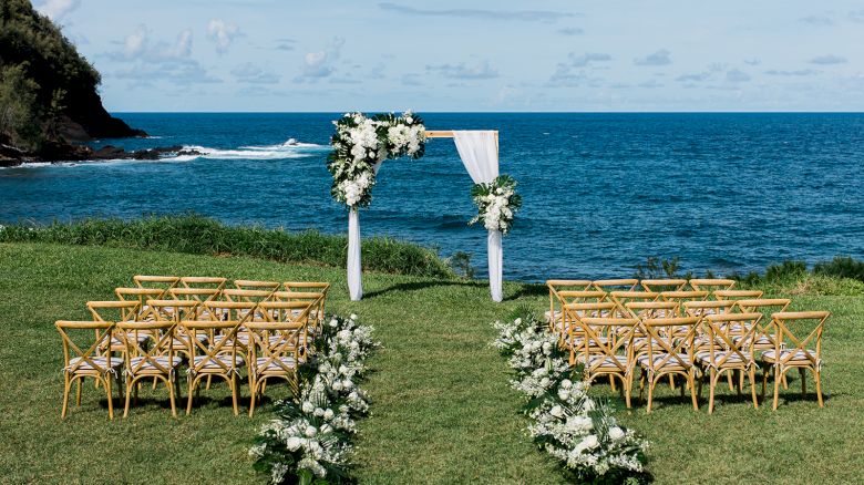 A seaside outdoor wedding setup with wooden chairs, a decorated arch with flowers and fabric, and floral arrangements lining the aisle.