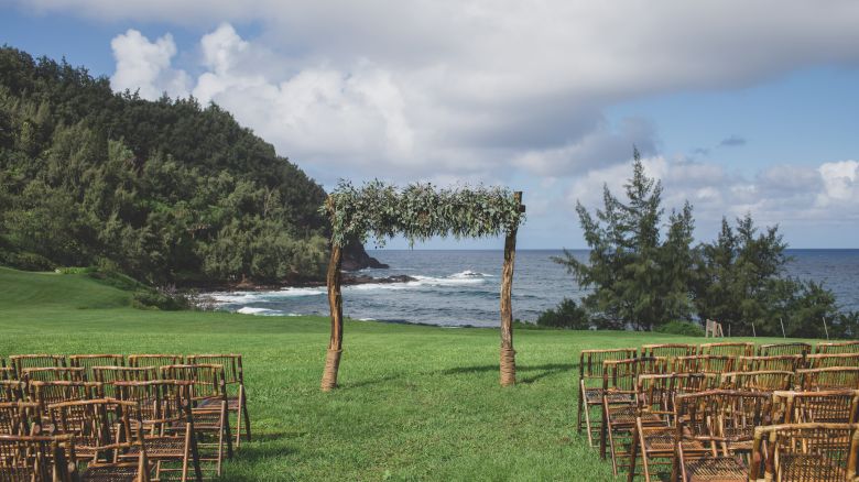 The image shows an outdoor wedding setup with wooden chairs and an arch decorated with greenery by the ocean and mountainous landscape.