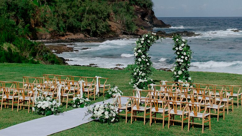 An outdoor wedding setup by the sea with wooden chairs, a white aisle, floral decorations, and a scenic backdrop of hills and the ocean.