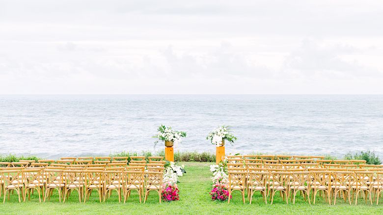 An outdoor wedding setup on a grassy area facing a body of water, with rows of wooden chairs and floral decorations.