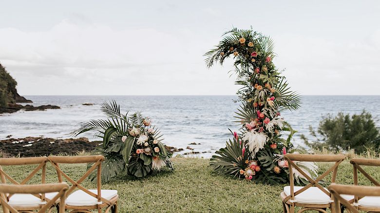 A seaside wedding setup with chairs facing an arch decorated with flowers and greenery, overlooking the ocean on a clear day.