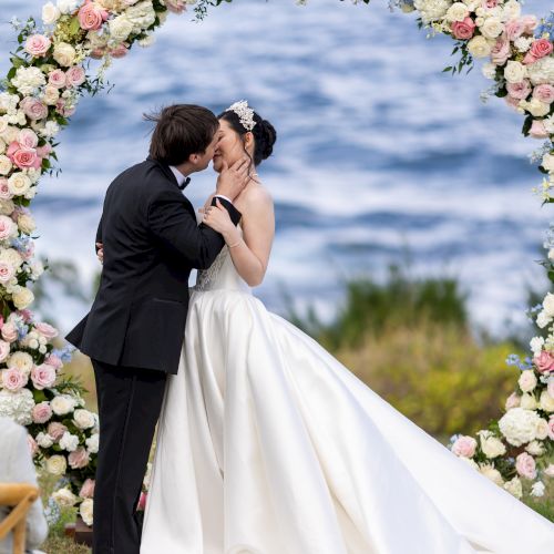 A couple is sharing a kiss under a flower arch at an outdoor wedding by the sea, with waves visible in the background.