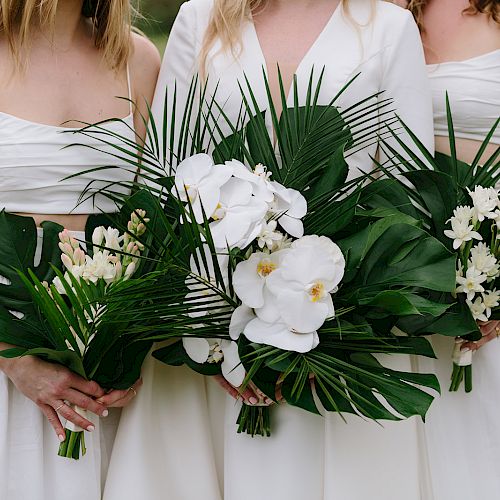 Three women in white dresses are holding bouquets made of green leaves and white flowers, including orchids.