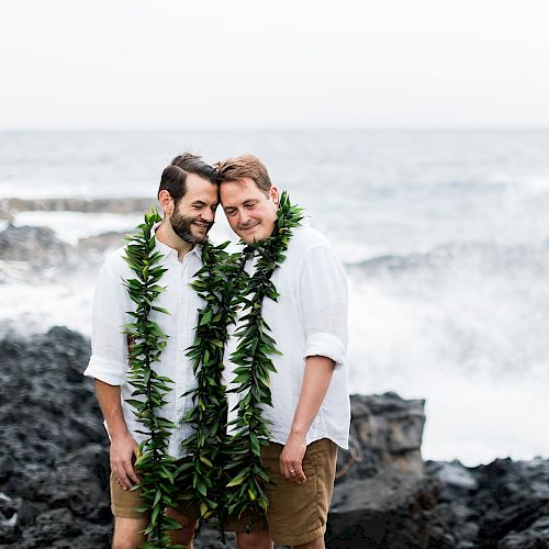 Two individuals wearing white shirts and green leaf garlands stand close together on a rocky shore with waves crashing behind them.