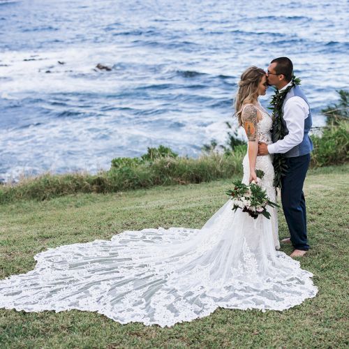 A couple shares a romantic kiss by the ocean, the bride's long train beautifully spread out on the grass, with scenic waves in the background.