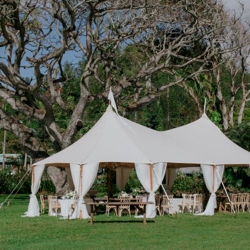 A white canopy tent is set up on a grassy area surrounded by trees, with tables and chairs arranged underneath, possibly for an outdoor event.