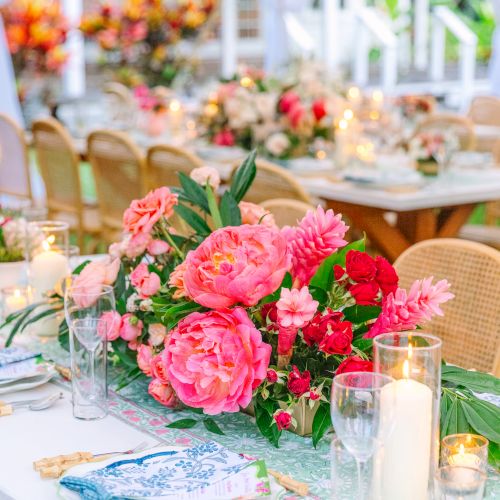 The image shows an elegantly set table in an outdoor setting with vibrant pink and red floral centerpieces, candles, and tableware.