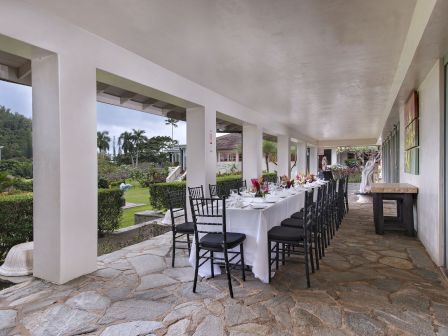 A long dining table set under a covered patio, surrounded by black chairs, with table settings and flower centerpieces, overlooking a garden.