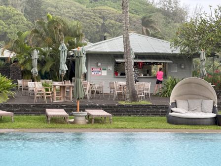 The image shows a poolside bar with tables, chairs, umbrellas, and a lounge area. Lush greenery surrounds the area, creating a tropical ambiance.