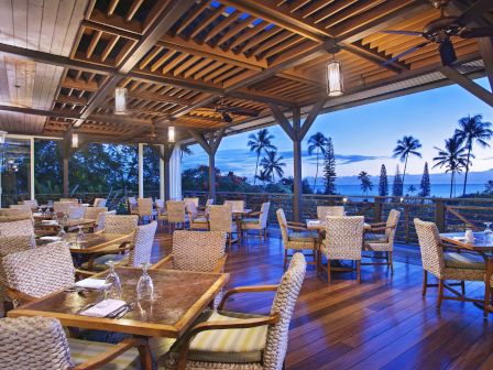 An open-air restaurant with wood flooring, wicker chairs, and set tables, overlooking a scenic view of palm trees and a serene blue sky during twilight.