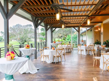 An outdoor event space with wooden flooring, draped tables, flower arrangements, and chairs, set up under a pergola with hanging lights.