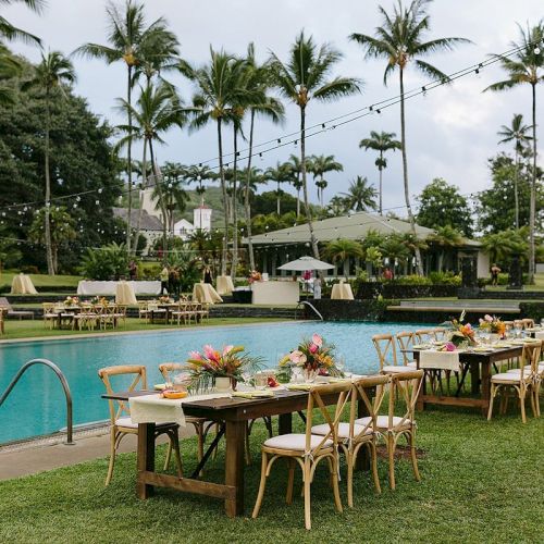 An outdoor dining setup beside a pool with decorated tables and chairs, surrounded by palm trees under a cloudy sky.