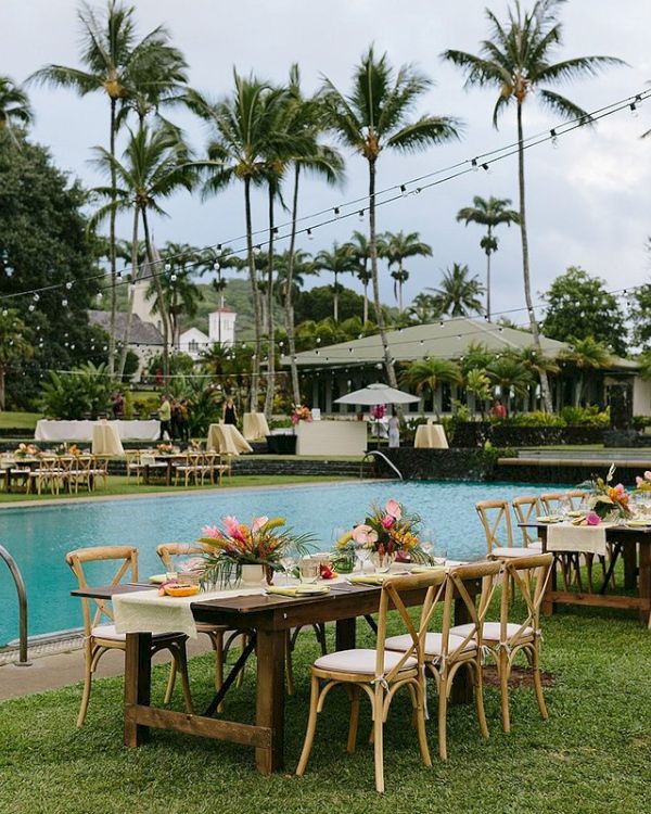 An outdoor dining setup beside a pool with decorated tables and chairs, surrounded by palm trees under a cloudy sky.