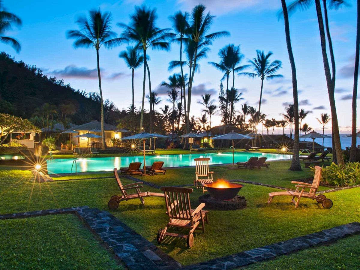 This image shows an outdoor setting with a pool, palm trees, and a group of chairs around a fire pit, likely at a resort during twilight.