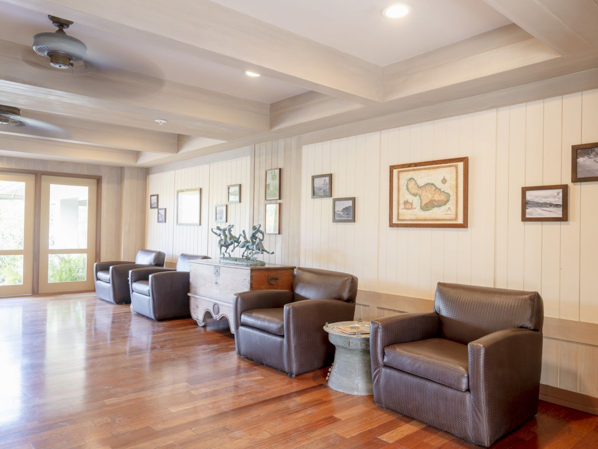 The image shows a room with wooden flooring, leather chairs, framed artwork on the walls, and a large window in the background.