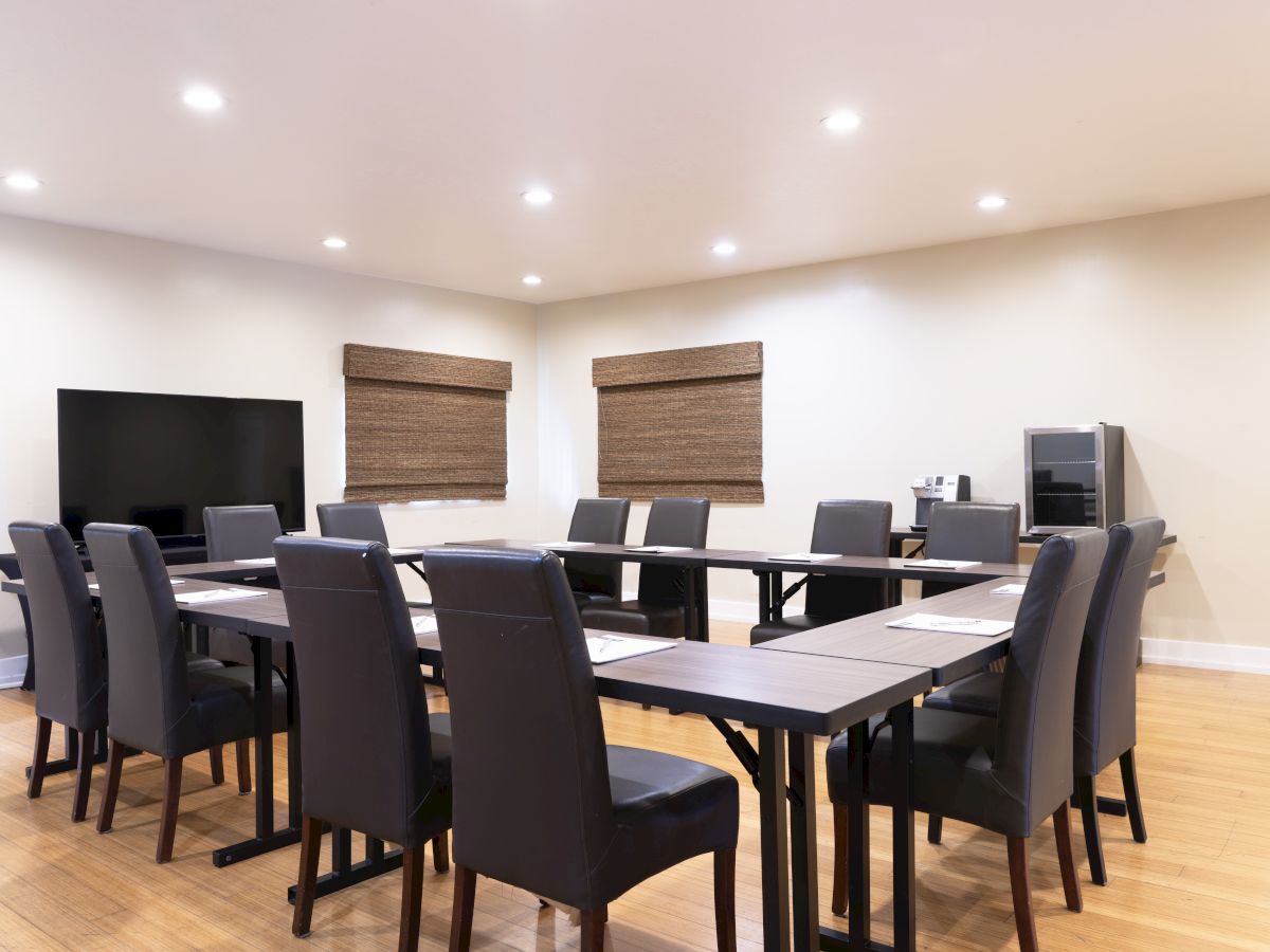 A modern conference room with a long table, several chairs, a TV, and windows with blinds on beige walls.