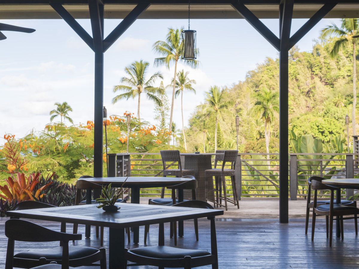 An outdoor seating area with tables and chairs, overlooking lush greenery and palm trees under a partly cloudy sky.