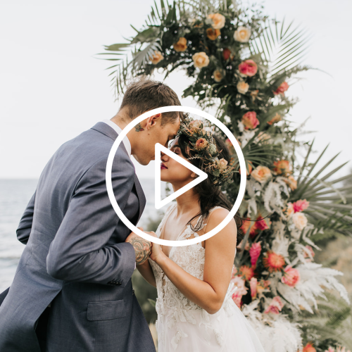 A couple is standing close together, dressed in wedding attire, in front of a flower arch by the sea. There is a play button overlay.