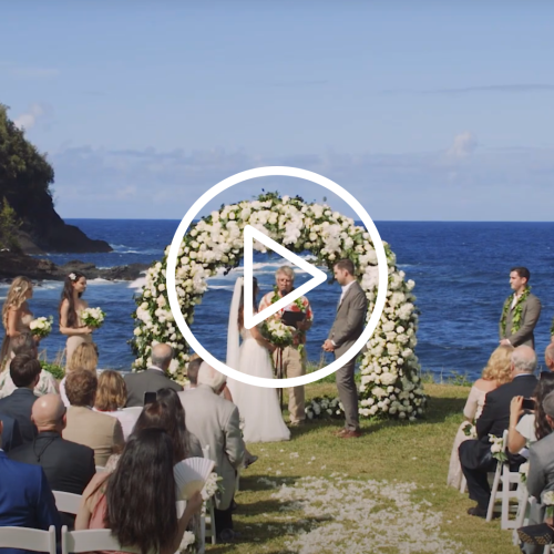 A wedding ceremony by the sea, with the couple standing under a flower arch, guests seated, and a scenic ocean backdrop under a clear sky.