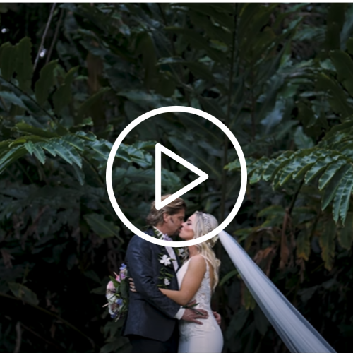 A couple is kissing in a lush, green, outdoor setting with a large play button overlay at the center, indicating a video.