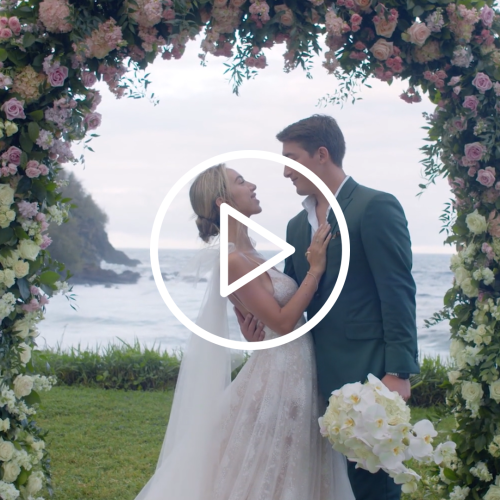 A couple stands under a flower arch by the ocean, dressed in wedding attire, holding a bouquet and looking at each other lovingly.