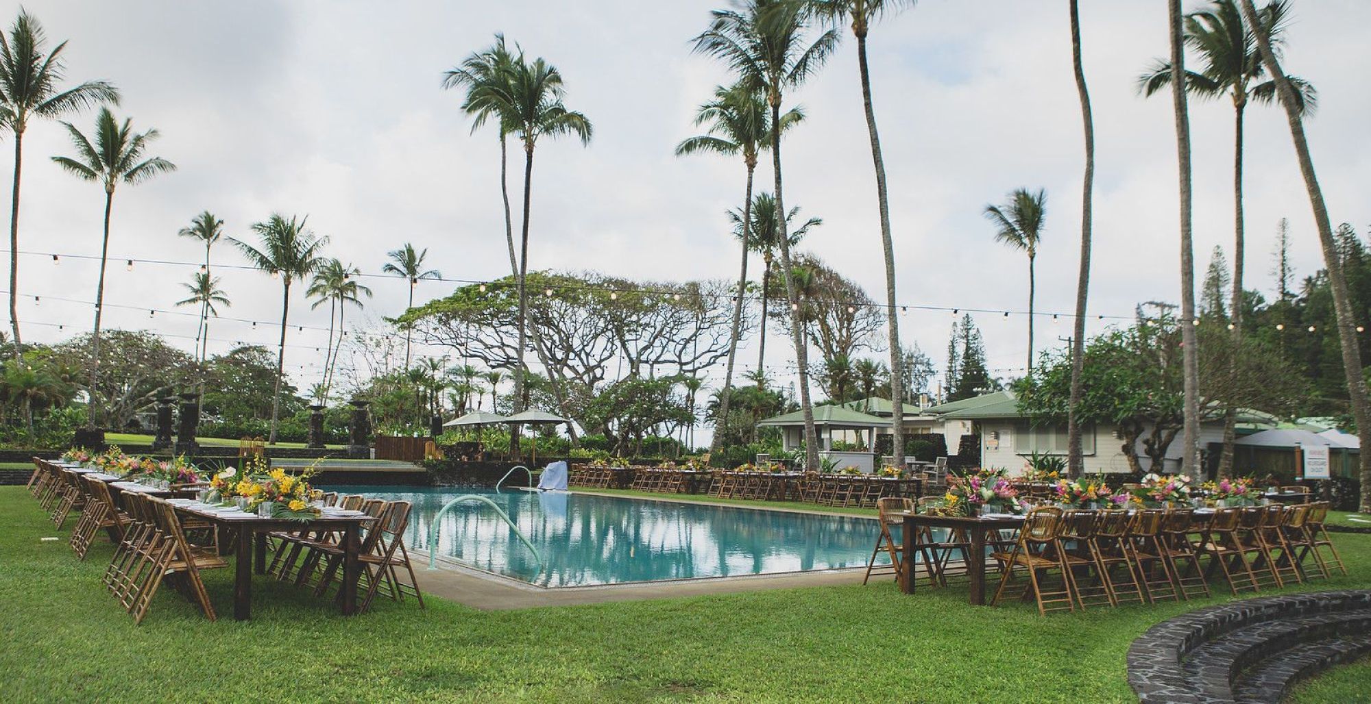 This image shows a tropical outdoor setting with tables and chairs arranged around a swimming pool, decorated with flowers and string lights.