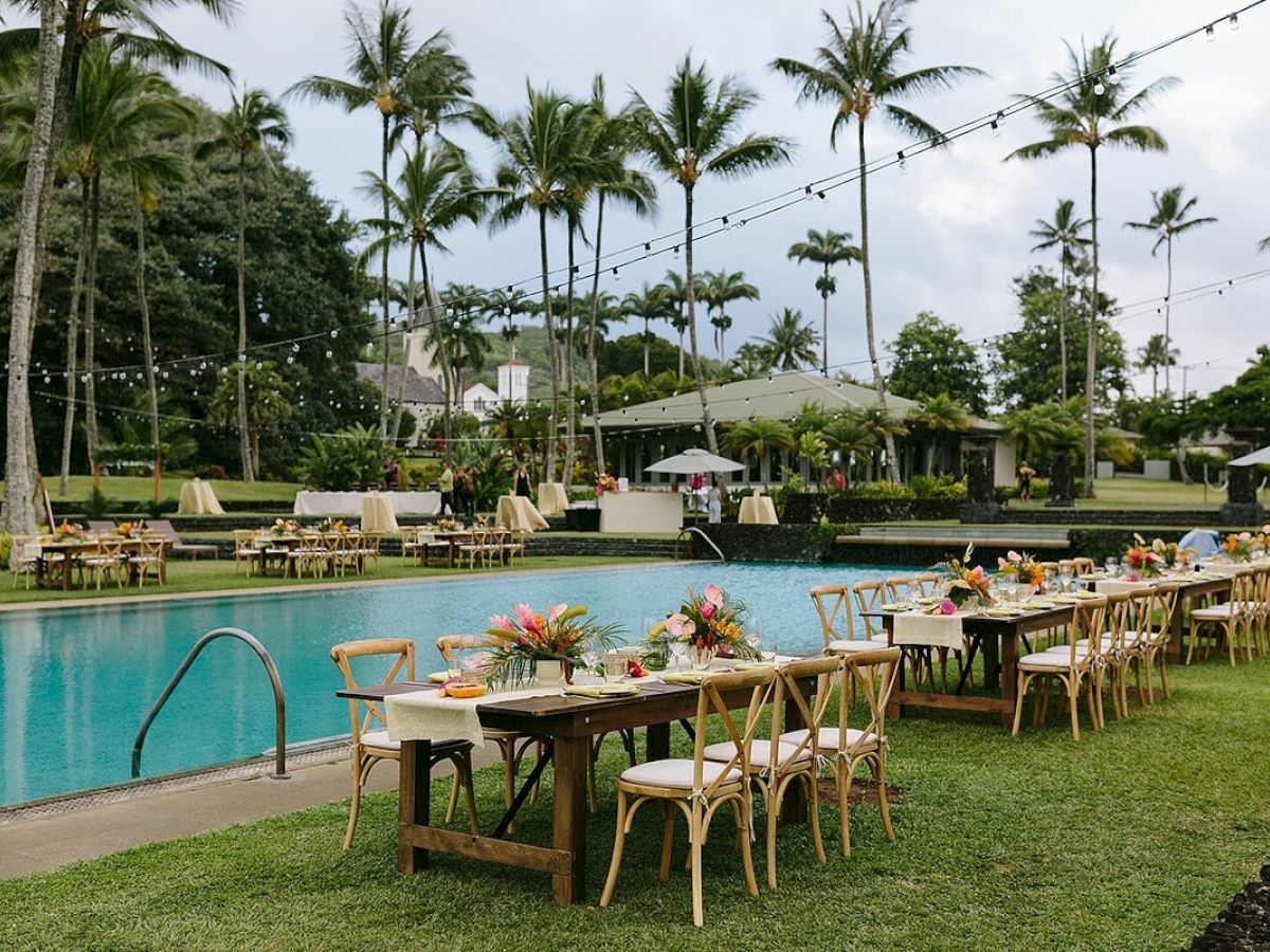 Outdoor poolside setup with tables and chairs, decorated with flowers, surrounded by palm trees and string lights, creating a tropical ambiance.