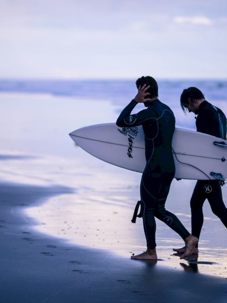 Two people in wetsuits are walking along the beach, one carrying a surfboard, with the ocean and a distant shore visible in the background.