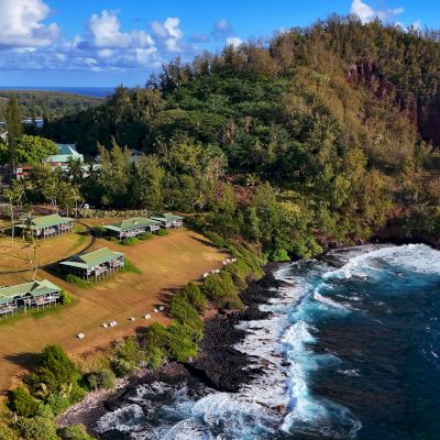 A coastal landscape with buildings, lush greenery, and waves crashing against the rocky shore under a partly cloudy sky.