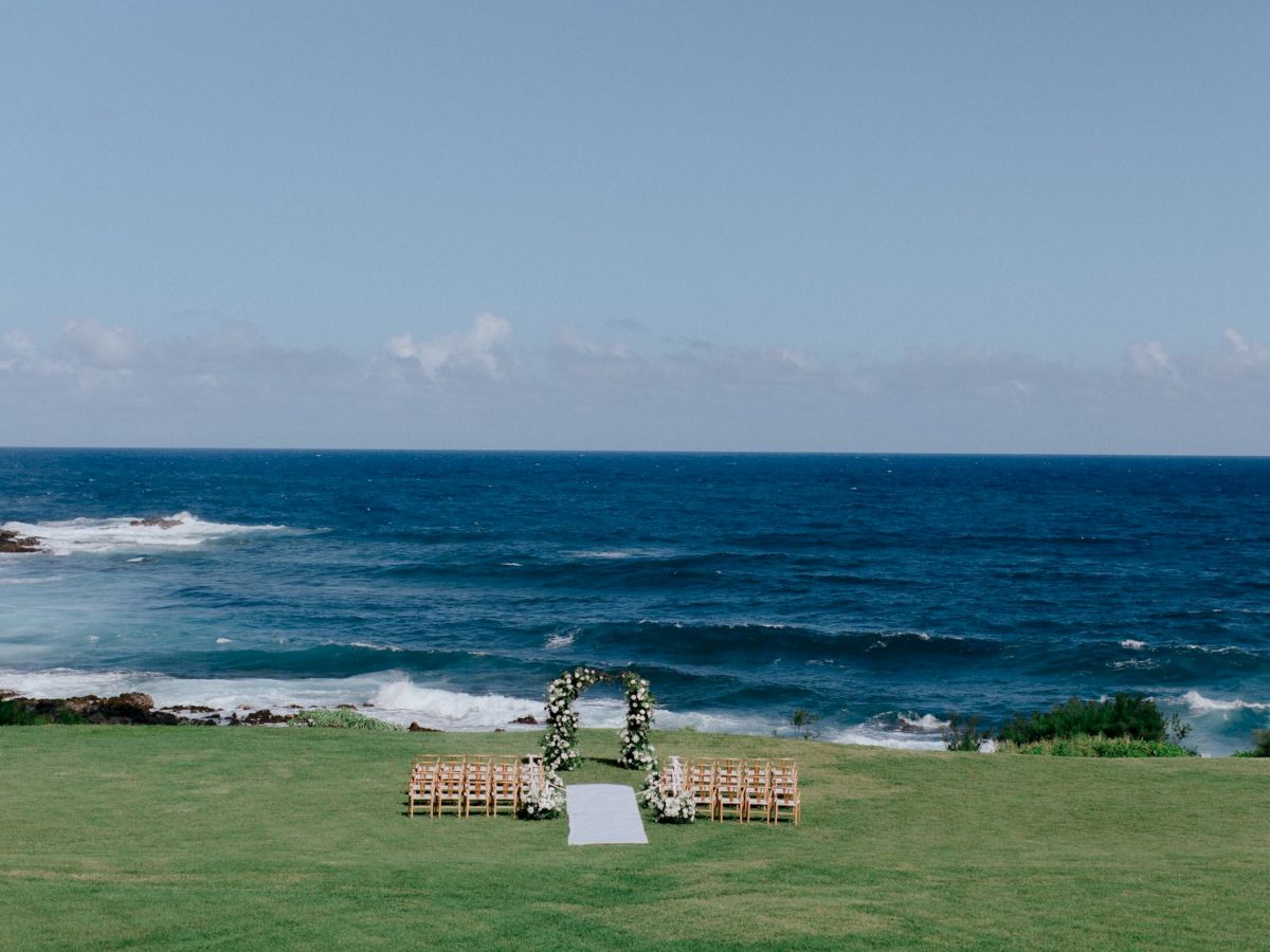 An outdoor wedding setup with a floral arch and seating on a grassy area, overlooking the sea, with waves crashing against the shore.