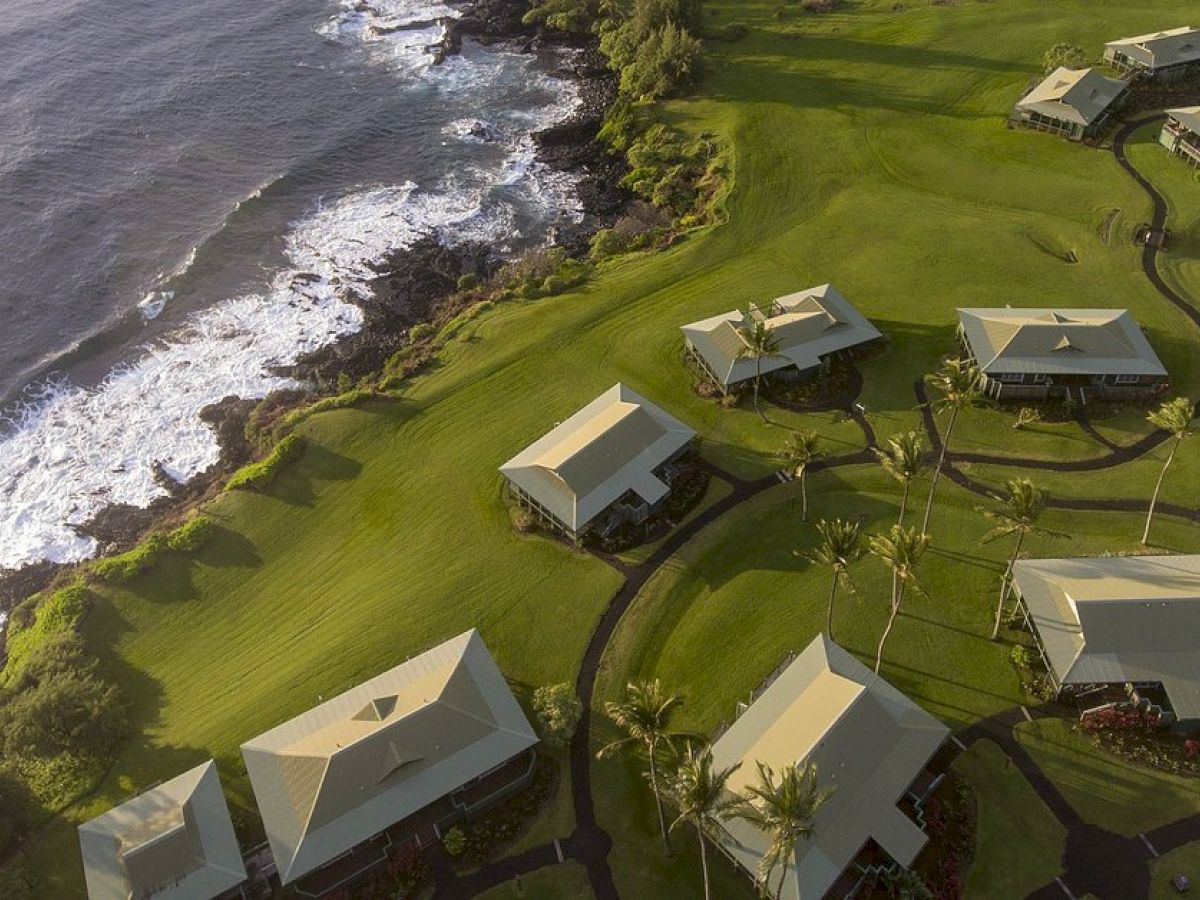The image features an aerial view of green coastal land with a cluster of buildings, pathways, and the ocean visible.