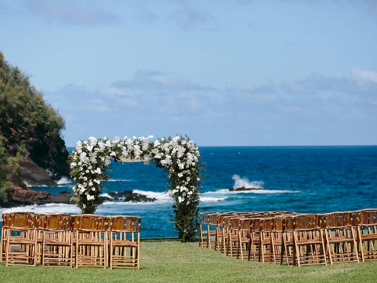 An outdoor wedding setup by the ocean, with wooden chairs arranged in rows and a floral arch overlooking the sea and a rocky cliff.