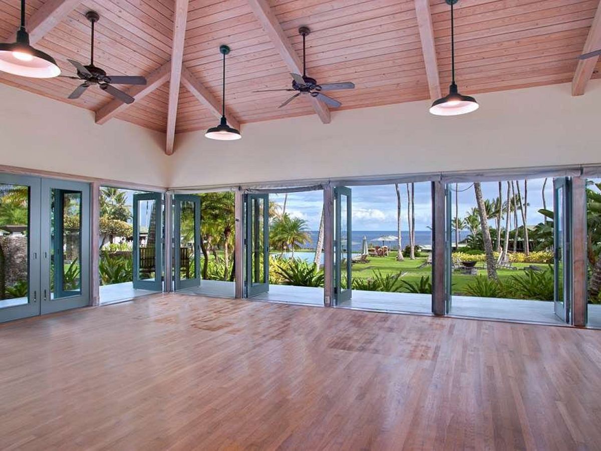 The image shows a spacious, empty room with wooden floors, a vaulted ceiling, multiple ceiling fans, and large windows offering a view of a tropical landscape.
