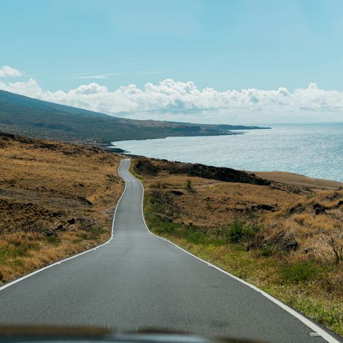 A coastal road stretches forward, flanked by dry, grassy fields on one side and the ocean on the other, under a clear blue sky with scattered clouds.