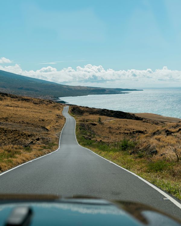 A coastal road stretches forward, flanked by dry, grassy fields on one side and the ocean on the other, under a clear blue sky with scattered clouds.