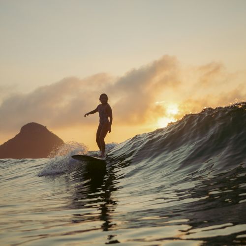 A person is surfing on a wave during sunset with mountains and clouds in the background, creating a serene and picturesque scene.