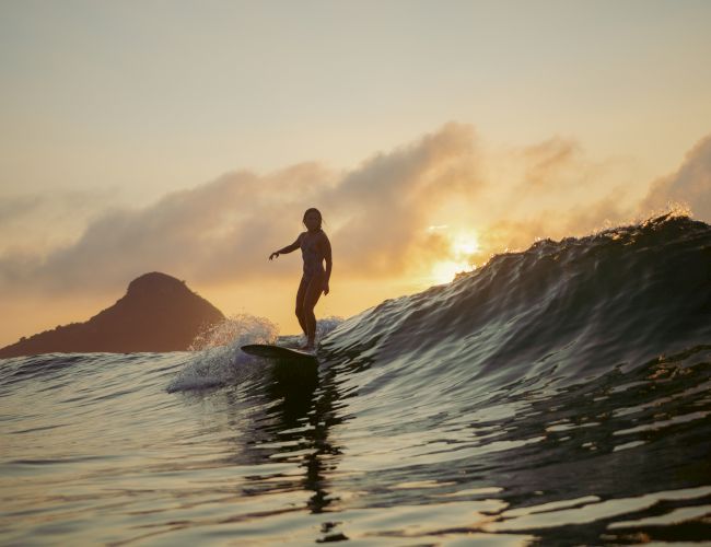 A person is surfing on a wave during sunset with mountains and clouds in the background, creating a serene and picturesque scene.