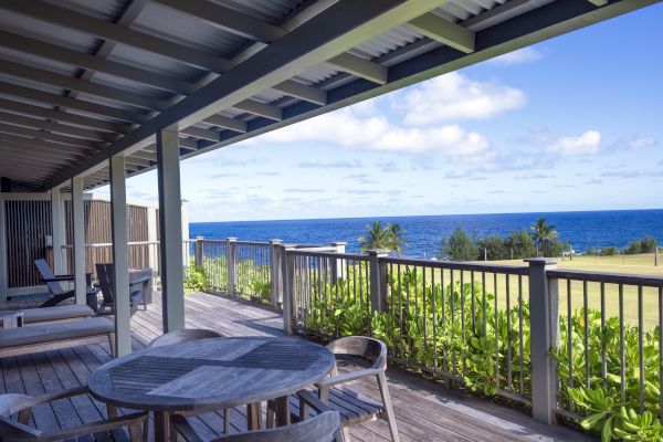 A wooden deck with chairs and tables overlooks a scenic ocean view under a blue sky with clouds.