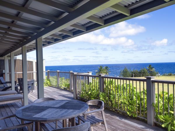 A wooden deck with chairs and tables overlooks a scenic ocean view under a blue sky with clouds.